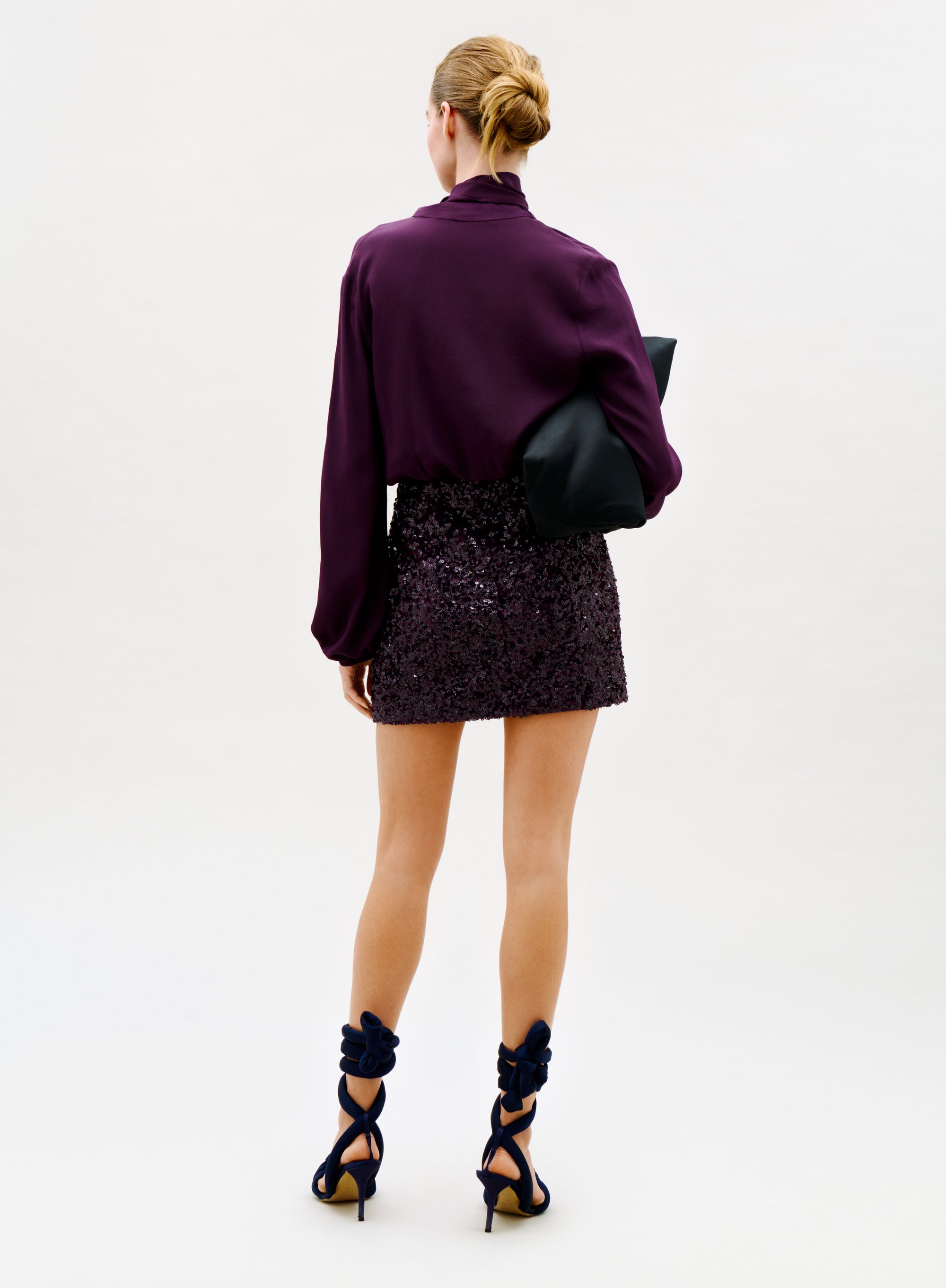 A woman with blonde hair in a neat bun, wearing the Faye Dress and black high heels with bows, holds a black clutch as she stands facing away against a plain light background.