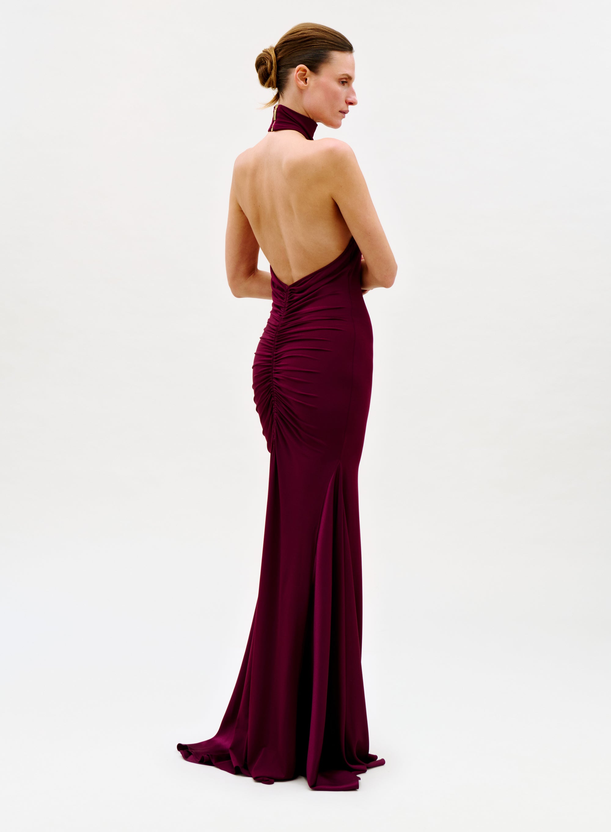A woman faces away, wearing the Feist Dress—a sleeveless, backless, floor-length burgundy gown with ruched back detailing. Her hair is in a neat bun against a plain white background.