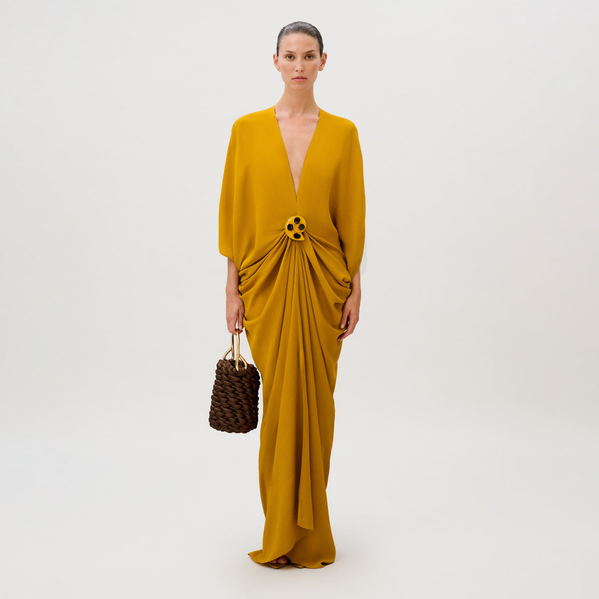 A model wears the floor-length, mustard yellow viscose Fiore Tunic featuring a V-neck, gathered waist, and dark decorative clasp, carrying a small, brown braided handbag.