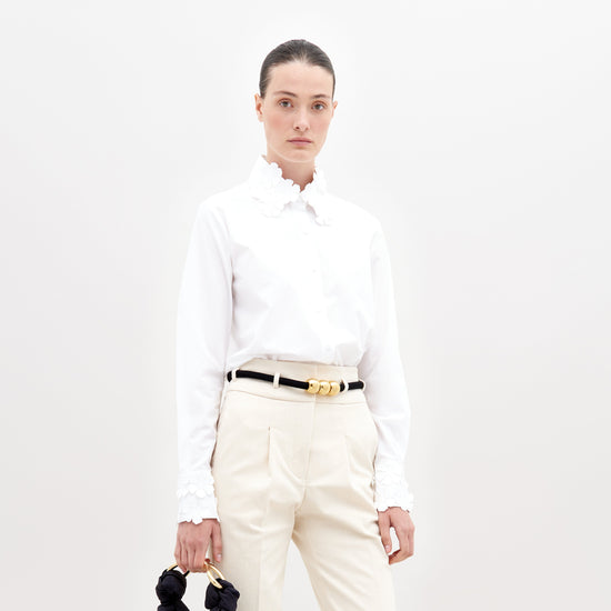 A person with dark hair tied back, wearing a Flower Blouse White with flower details on the collar and sleeves, beige pants, and a black belt with gold accents, holding a small black handbag. Pre-order now to ensure your outfit ships by November 15th, 2024.