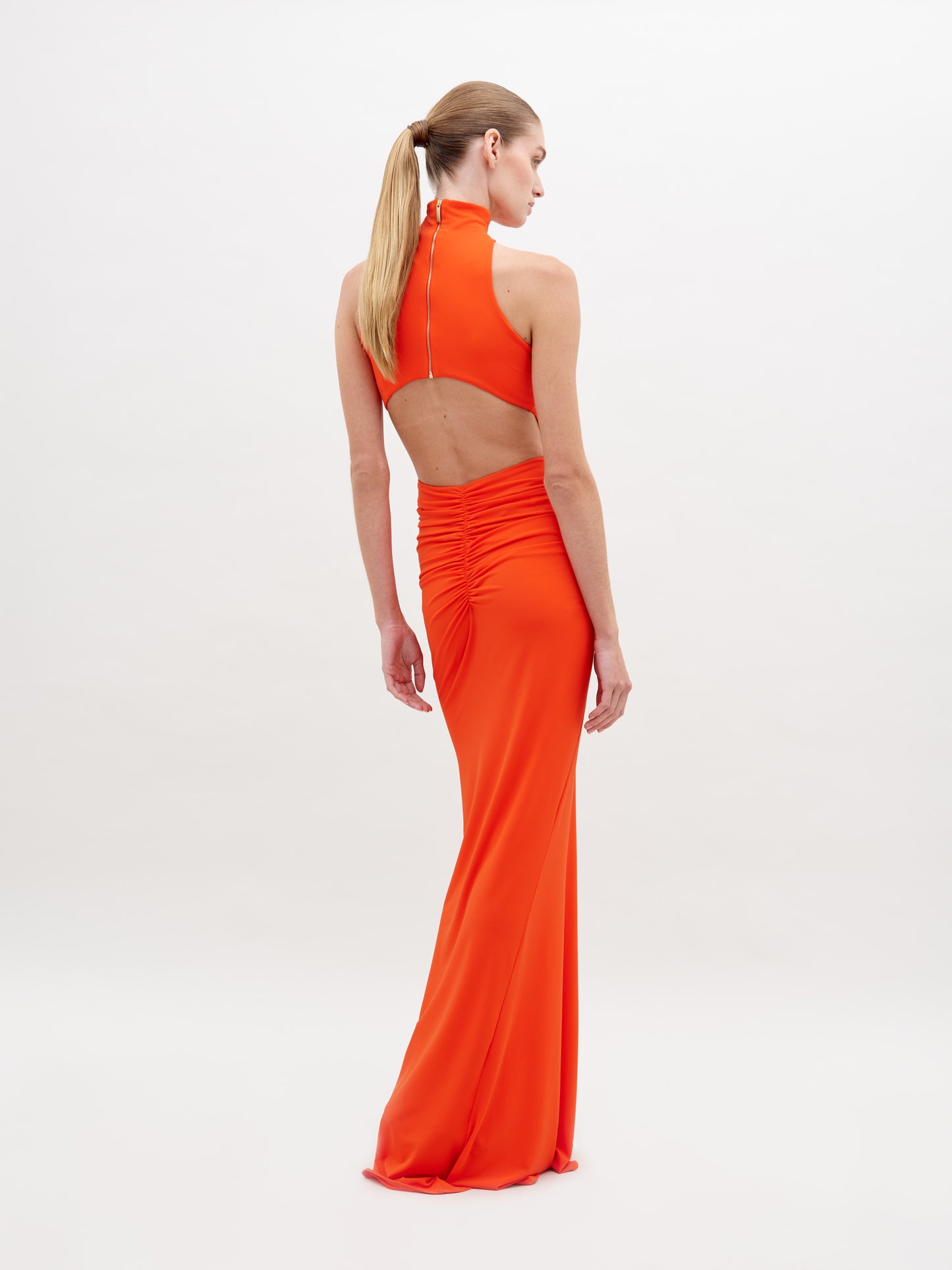 A woman in a long, sleeveless, high-neck orange Frances Dress Tangerine stands against a plain white background. The dress is gathered at the waist with a decorative clasp. This stylish piece is available for pre-order now and will ship by November 15th, 2024.