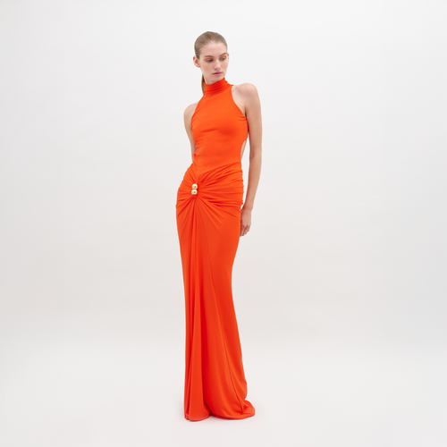 A woman in a long, sleeveless, high-neck orange Frances Dress Tangerine stands against a plain white background. The dress is gathered at the waist with a decorative clasp. This stylish piece is available for pre-order now and will ship by November 15th, 2024.