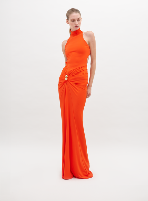 A woman in a long, sleeveless, high-neck orange Frances Dress Tangerine stands against a plain white background. The dress is gathered at the waist with a decorative clasp. This stylish piece is available for pre-order now and will ship by November 15th, 2024.