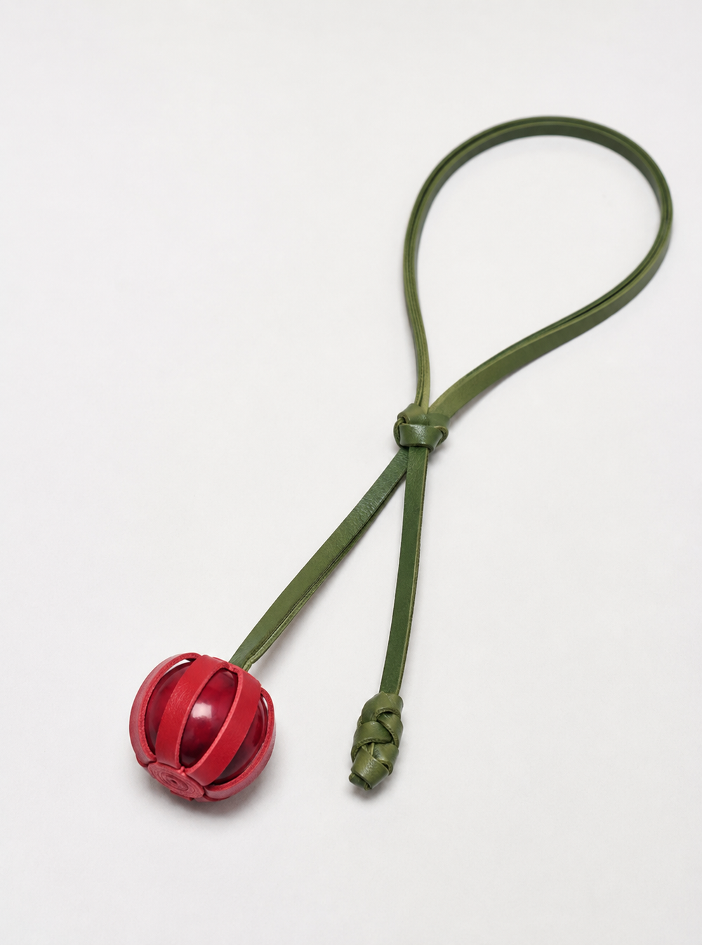 The Fruit Necklace features a red leather ball with open slits, attached to a green leather looped strap, displayed on a plain white background.