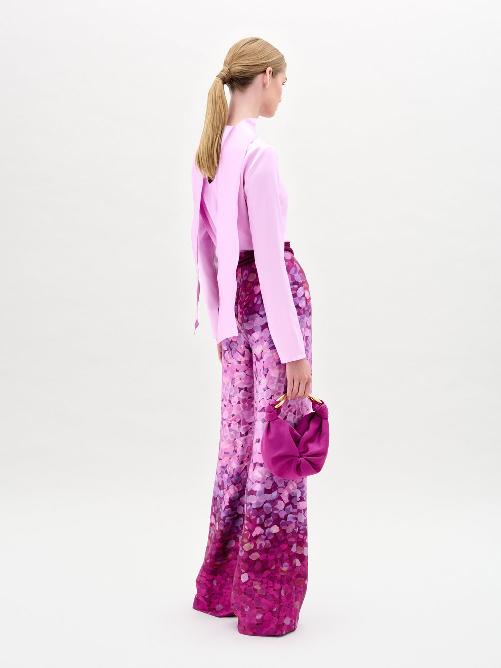 A woman stands facing forward, wearing a light pink long-sleeve top, Como Pant in high-waisted Italian fabric with pink and purple patterns, and holding a matching pink handbag.