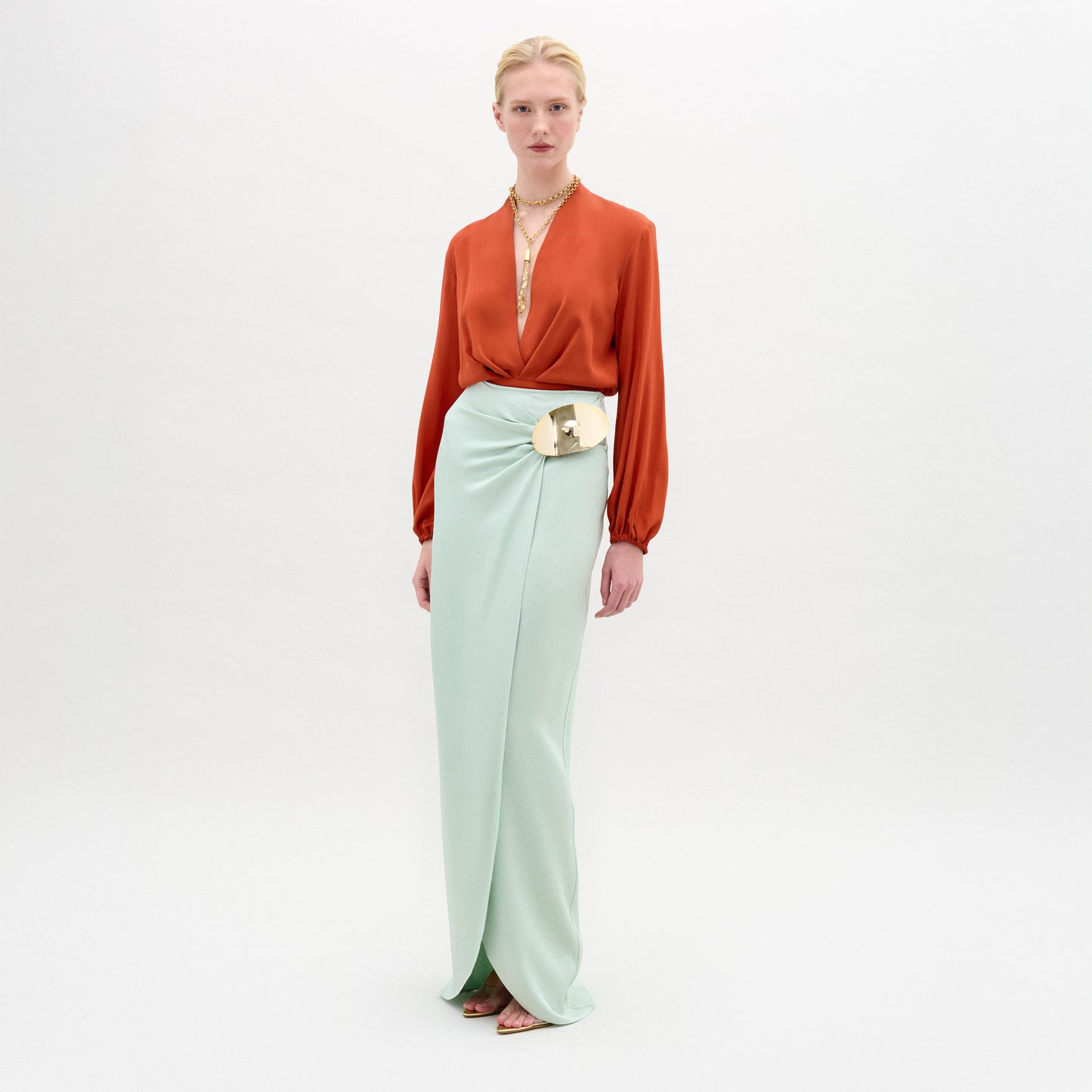 A person stands against a plain white background wearing the Gadea Skirt in light blue Italian fabric, an orange long-sleeve blouse, a gold belt, and a necklace.