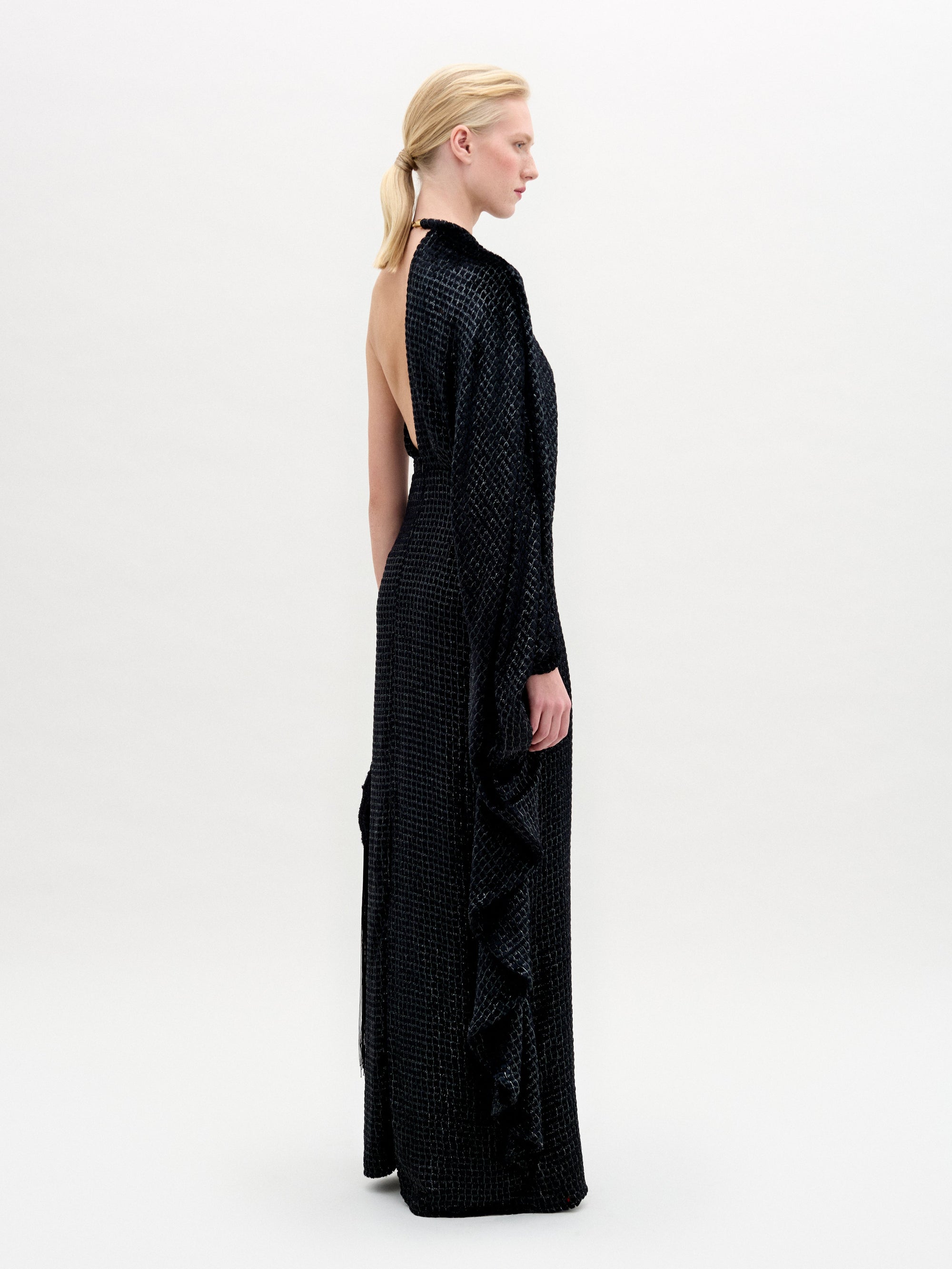 A woman stands against a plain background wearing the Gael Dress—a black, floor-length maxi with an asymmetrical neckline and textured pattern—holding a small clutch in her right hand.