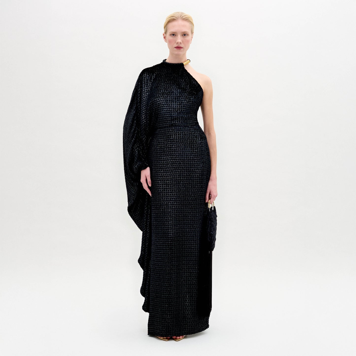 A woman stands against a plain background wearing the Gael Dress—a black, floor-length maxi with an asymmetrical neckline and textured pattern—holding a small clutch in her right hand.