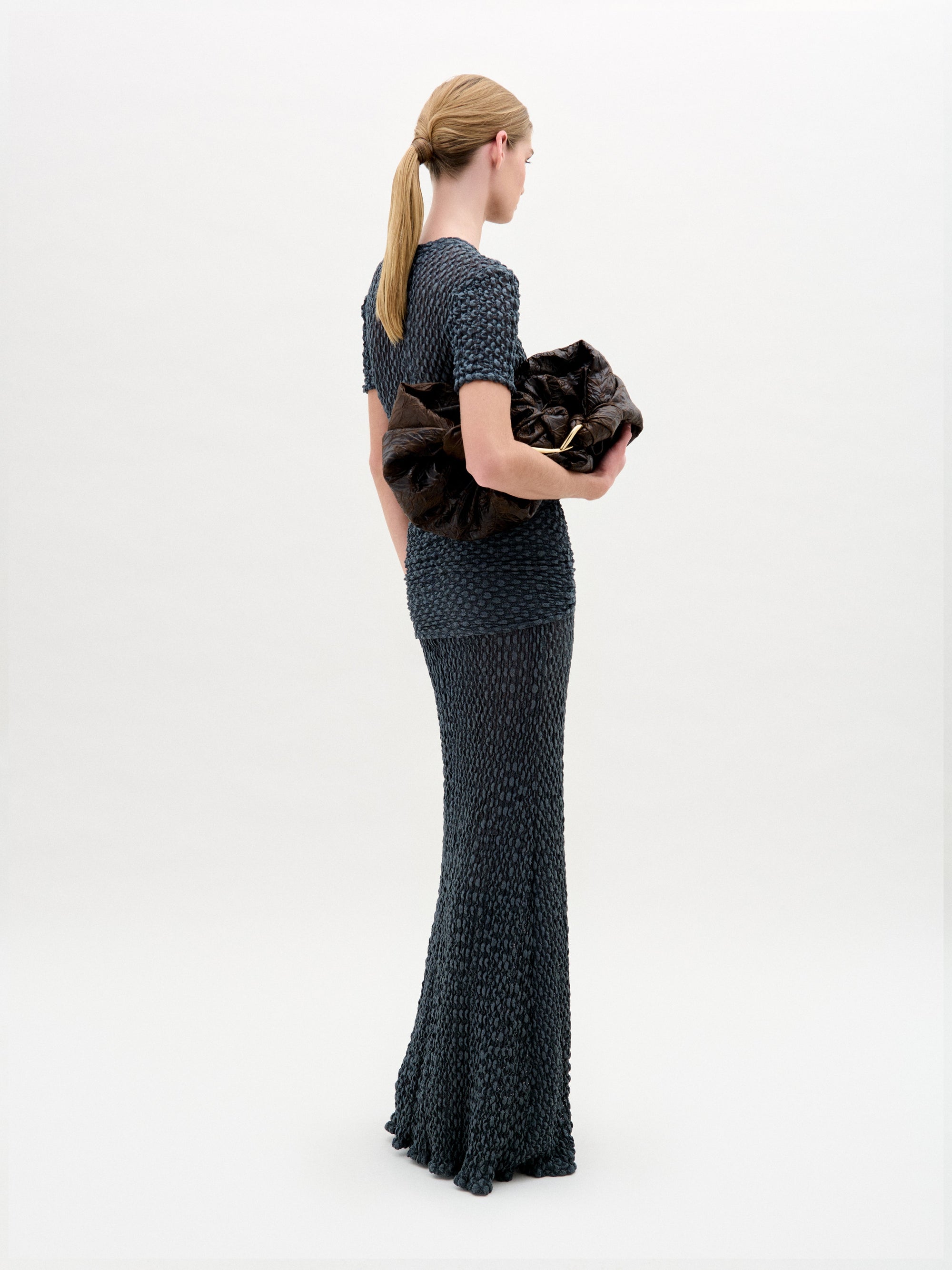 A woman with blonde hair in a low ponytail wears the Gen Tshirt, a textured, fitted, dark short-sleeve top with knot detail, and holds a large dark ruched bag against a plain white background.