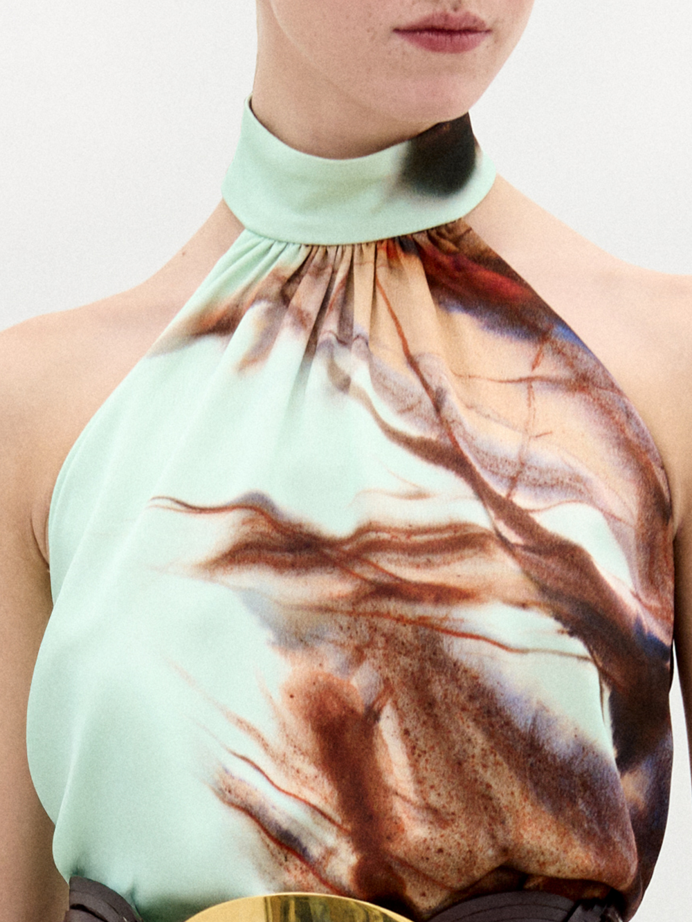 A woman stands against a plain white background wearing the Geovanna Blouse—a sleeveless, high-neck halter top featuring a brown and light blue abstract pattern—paired with a wide belt and matching skirt, made in Colombia.