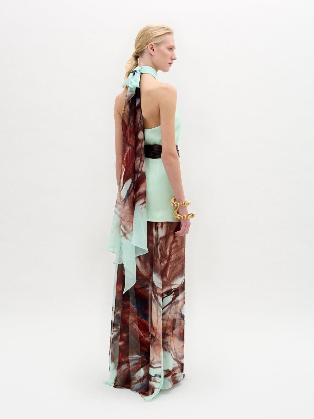 A woman poses against a plain backdrop in the Amalia Skirt—a flowing maxi with an abstract brown and blue pattern—paired with a sleeveless halter top, wide black belt, and bracelets. The look features luxurious Italian fabric.