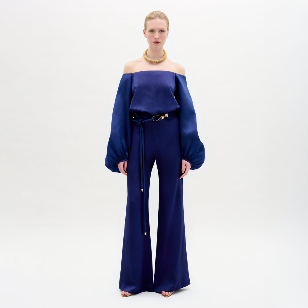 A Person wearing the Geraldine Jumpsuit in color Navy with a golden accesories on a white background.