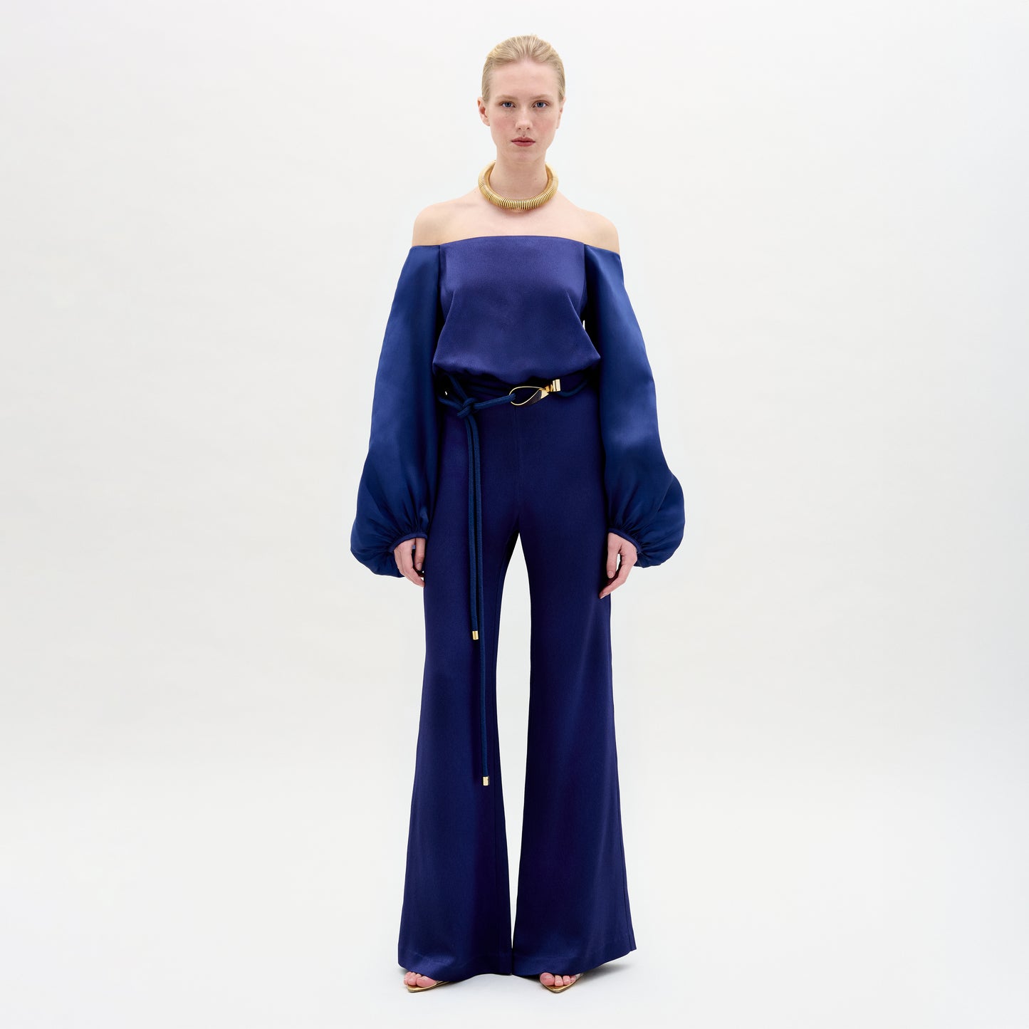 A Person wearing the Geraldine Jumpsuit in color Navy with a golden accesories on a white background.