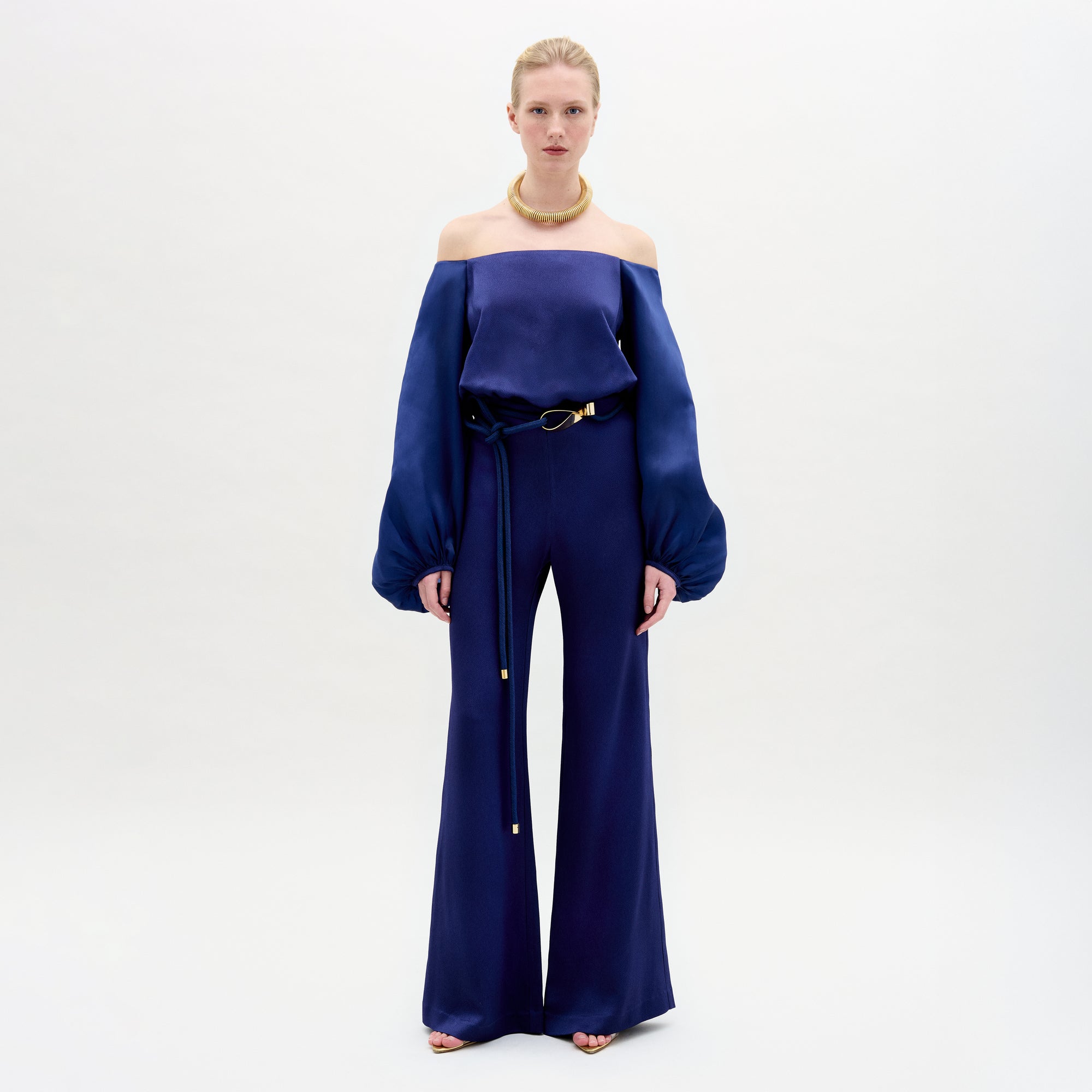 A Person wearing the Geraldine Jumpsuit in color Navy with a golden accesories on a white background.