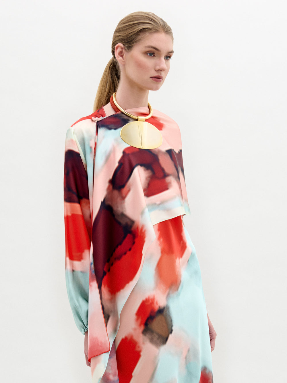 A woman in a colorful abstract-patterned dress accessorized with a circular gold necklace and the Gery Shrug stands against a plain background.