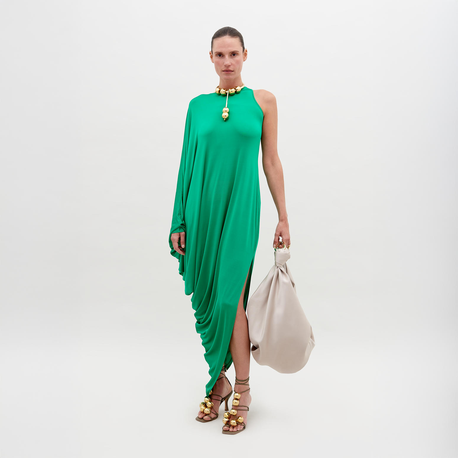 A person stands against a plain background, wearing a Golda Dress Green with gold accessories, holding a large beige handbag. Pre-order now for shipping by November 15th, 2024.