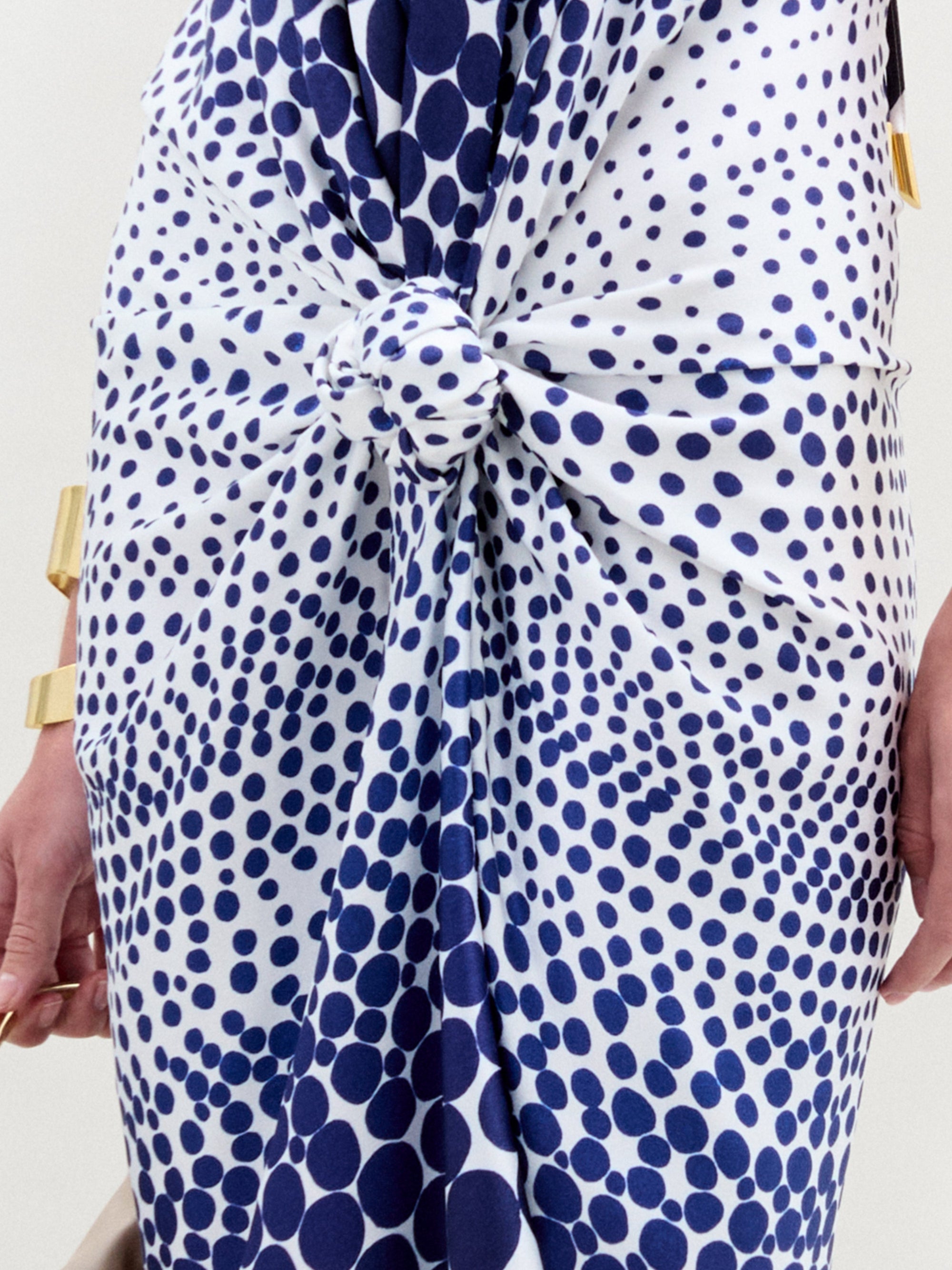 A woman in the Guadalupe Dress—a blue and white polka dot silk midi with a draped silhouette—pairs it with gold heels, gold cuff bracelets, and a beige handbag, standing against a plain white background.