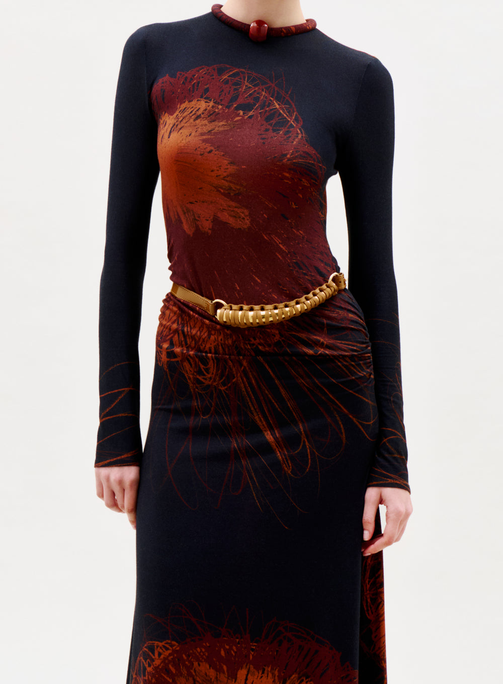 A person in the Gabbice Dress—a long-sleeved, fitted black dress with abstract red and brown patterns—paired with a chunky gold necklace and a gold belt worn diagonally, stands against a plain light background.