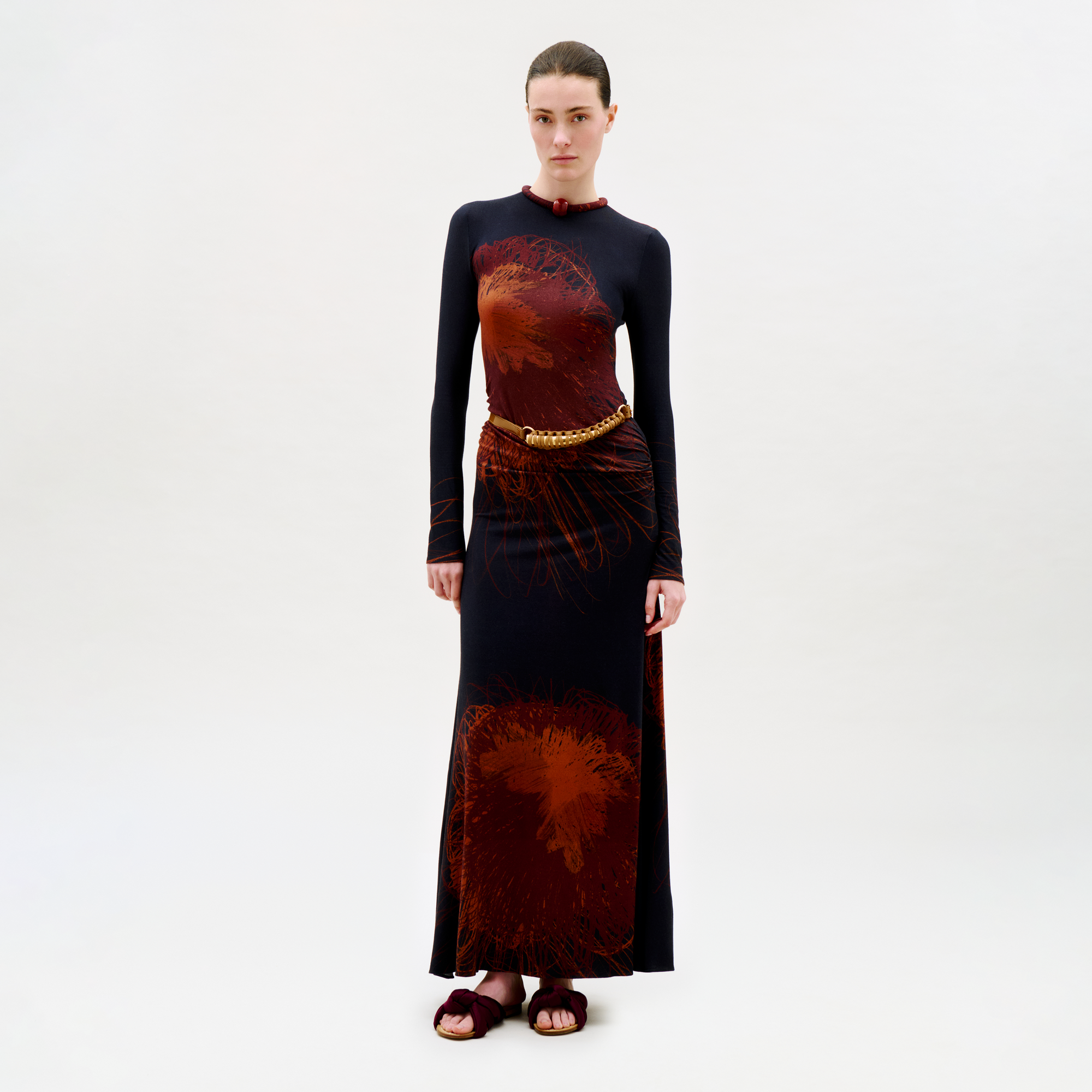 A woman with slicked-back hair and a neutral expression stands against a white background wearing the floor-length, long-sleeved Gabbice Dress in black with bold red floral patterns, styled with a braided belt and dark sandals.