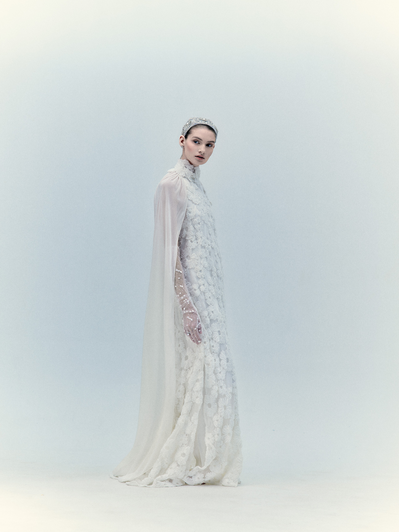 A woman in the Daniella Dress White, featuring a floor-length design and a unique cut-out in the center, stands against a plain background, looking downward. This exclusive piece highlights elegance and sophistication—available upon request.