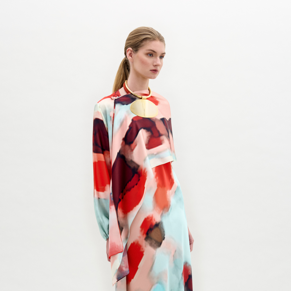 A woman in a colorful abstract-patterned dress accessorized with a circular gold necklace and the Gery Shrug stands against a plain background.