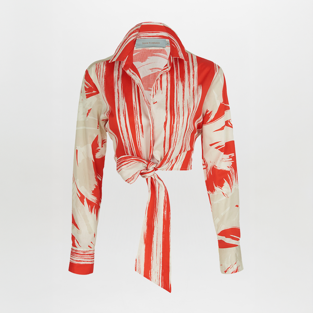 The Gisela Blouse Coral Red Palm Print by Silvia Tcherassi showcases a long-sleeve, button-up design with coral red palm print fabric, featuring abstract stripes and a stylish collar with a tied knot at the waist.