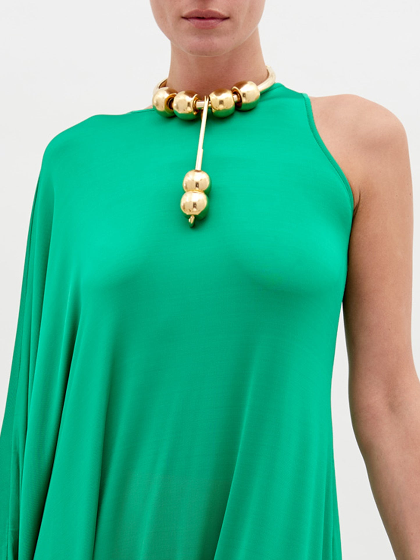 A person stands against a plain background, wearing a Golda Dress Green with gold accessories, holding a large beige handbag. Pre-order now for shipping by November 15th, 2024.