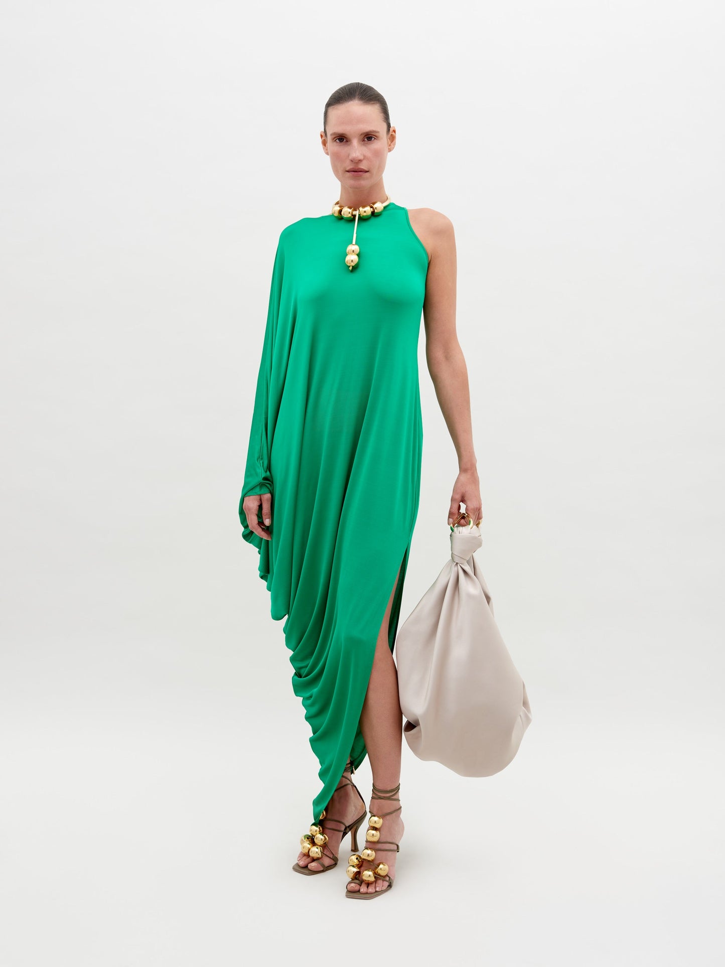 A person stands against a plain background, wearing a Golda Dress Green with gold accessories, holding a large beige handbag. Pre-order now for shipping by November 15th, 2024.