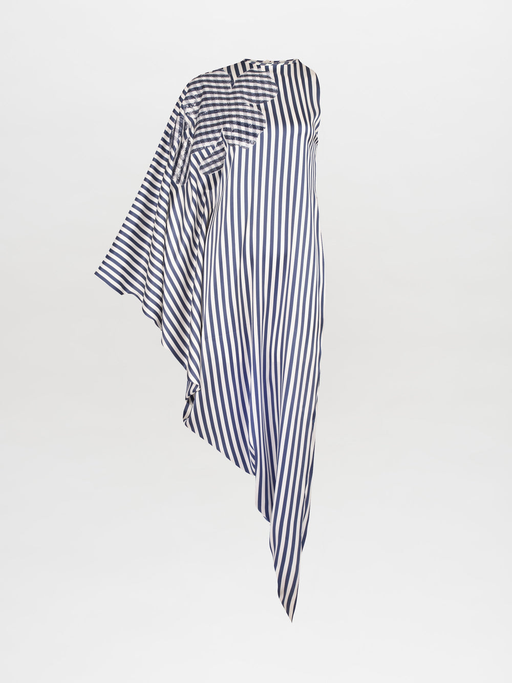 Person wearing the Golda Dress in White and Blue Stripes, an asymmetrical design with draped fabric, paired with white strappy sandals featuring embellishments, standing against a plain white background. Pre-order now to ensure it ships by February 15th, 2025.