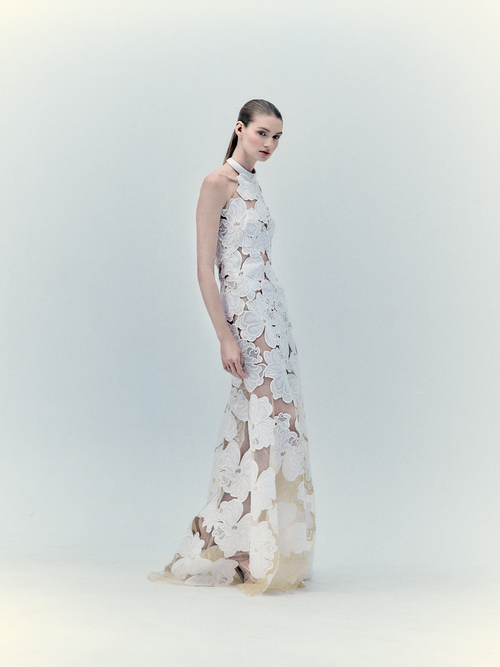 A model wearing the Grace Dress Pearl, an exclusive sleeveless white floral lace gown, posing against a light blue backdrop.