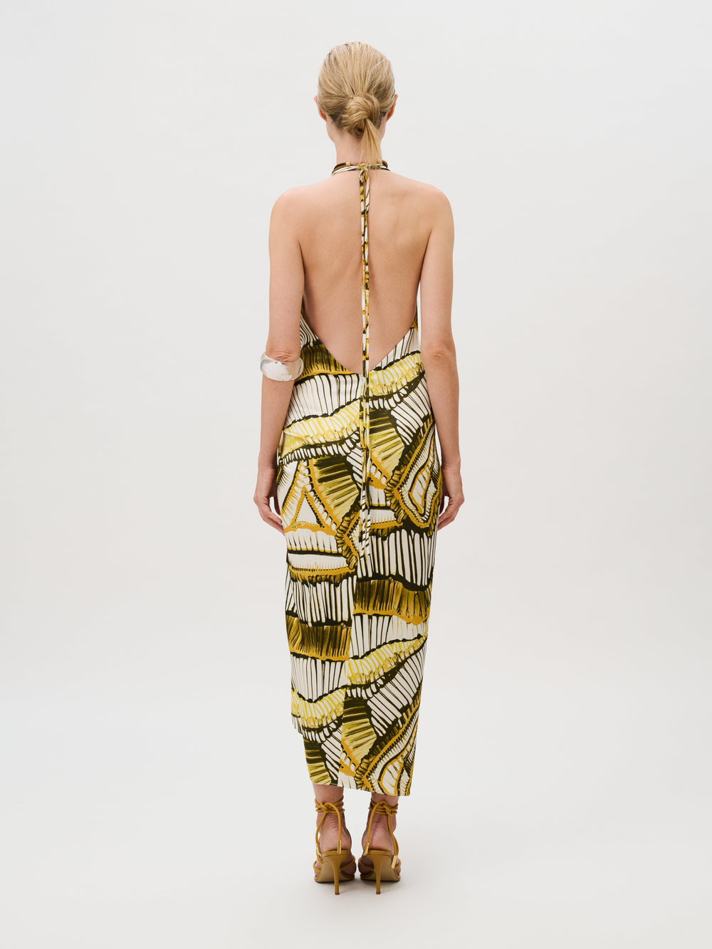 Back view of a person wearing the Guadalupe Dress, a sleeveless, backless midi dress of Italian fabric with a yellow and green geometric pattern, paired with strappy high-heeled sandals.