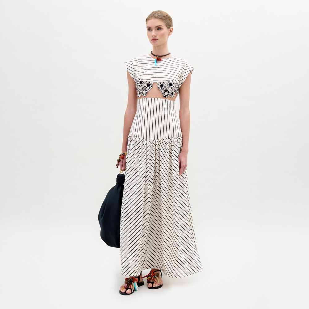 A woman stands against a white background wearing the Hanane Dress—a maxi dress with a cutout midsection—paired with black sandals, a necklace, and holding a large black bag.