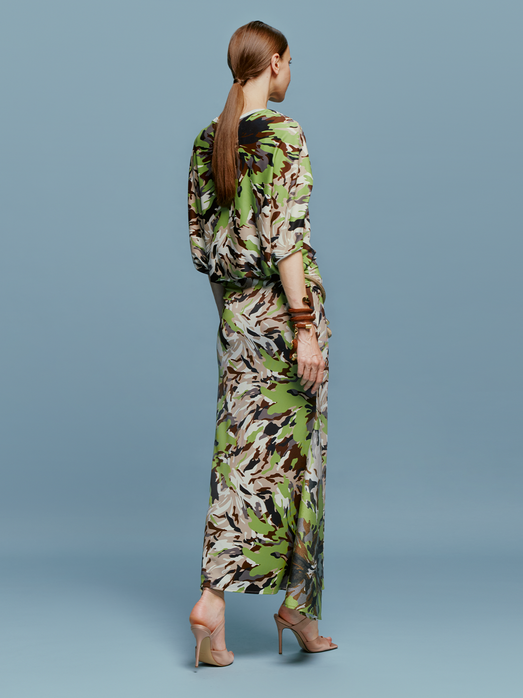 On August 19th, a woman poses against a light blue background wearing the Harriet Dress Floral Camo in green, black, and brown floral pattern. She is accessorized with high-heeled shoes and a bracelet.