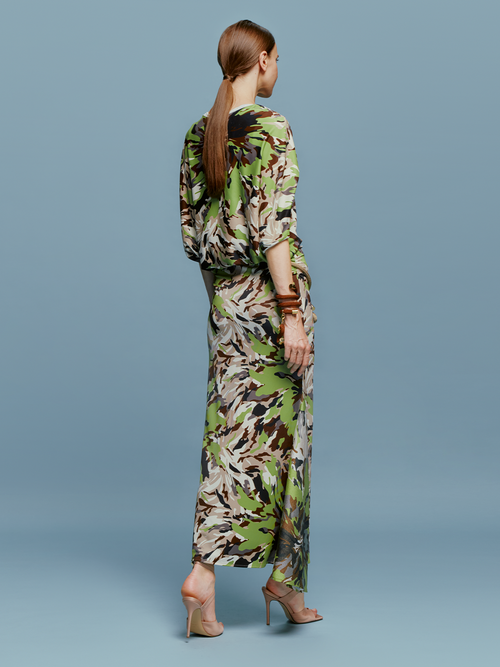 On August 19th, a woman poses against a light blue background wearing the Harriet Dress Floral Camo in green, black, and brown floral pattern. She is accessorized with high-heeled shoes and a bracelet.