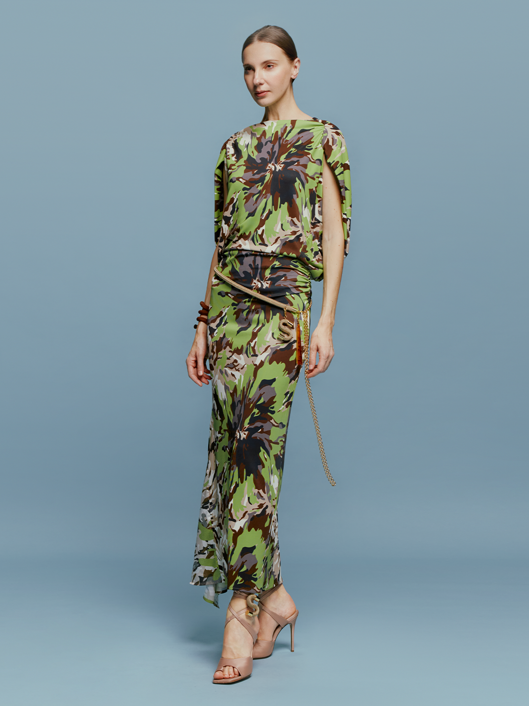 On August 19th, a woman poses against a light blue background wearing the Harriet Dress Floral Camo in green, black, and brown floral pattern. She is accessorized with high-heeled shoes and a bracelet.