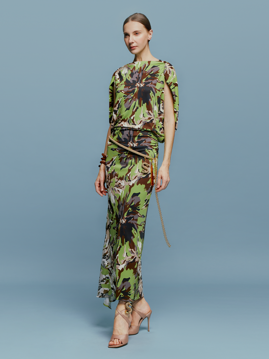 On August 19th, a woman poses against a light blue background wearing the Harriet Dress Floral Camo in green, black, and brown floral pattern. She is accessorized with high-heeled shoes and a bracelet.