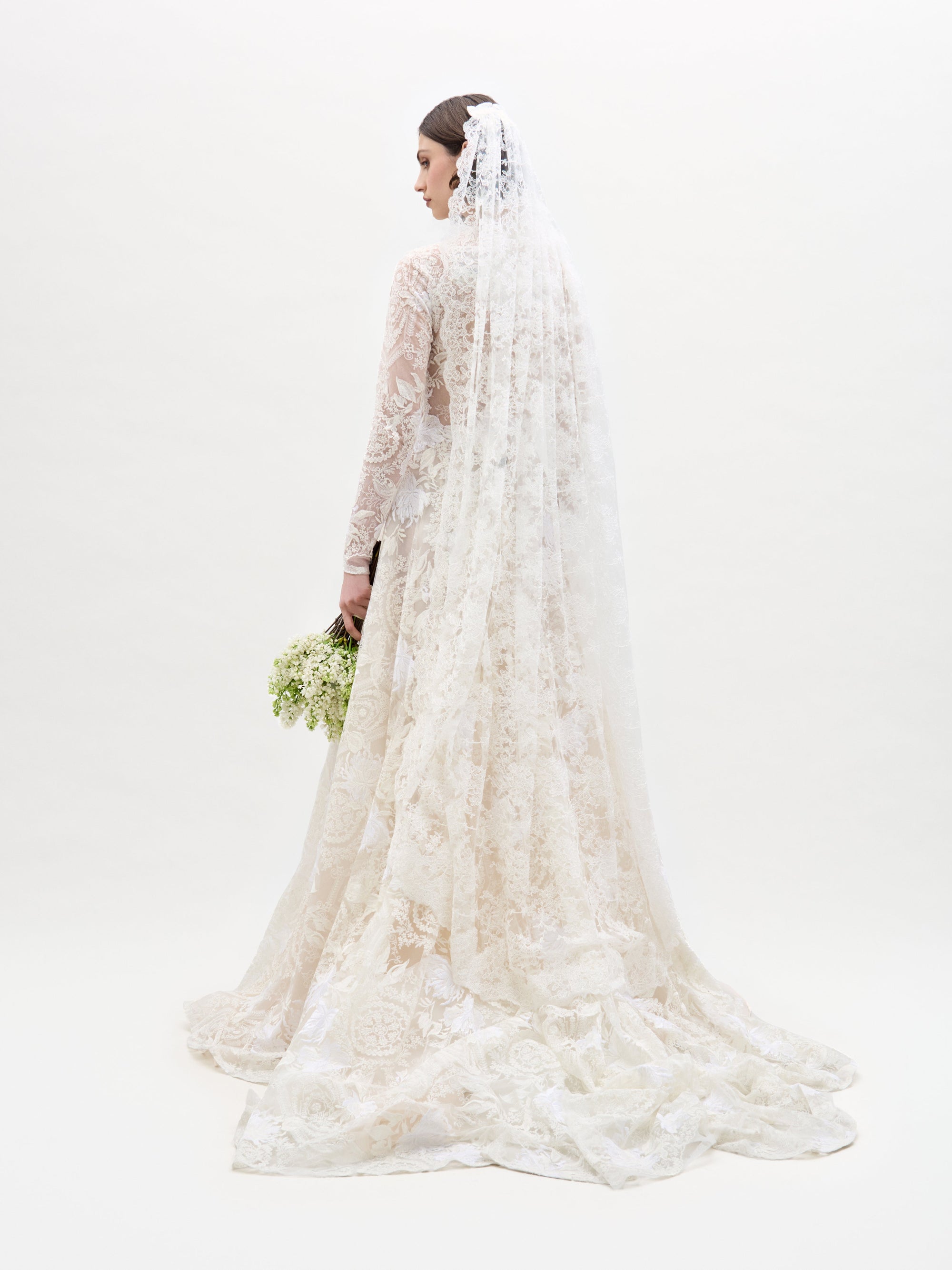 A woman poses against a plain background in the Hermoine Dress, an ornate lace bridal gown featuring long sleeves and a flowing train.