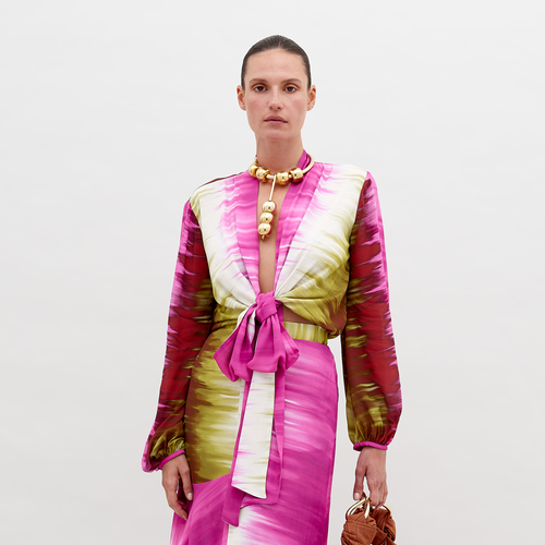 A woman with tied-back hair wears a Honey Blouse Violeta Lime Tie Dye with a front knot and matching skirt, accessorized with a gold bead necklace and holding a brown handbag against a plain background. This pre-order item will ship by November 15th, 2024.