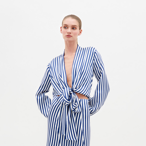 A person stands in the Honey Blouse White Blue Stripes, featuring a tie at the waist and a ruffled hem, paired with embellished sandals. This stylish ensemble is available for pre-order with shipping by February 15th.