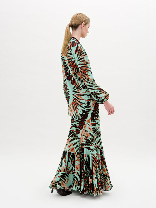 Against a plain white background, a person wears the Florina Skirt, crafted from luxurious Italian fabric. This long skirt showcases a vibrant tie-dye pattern with green, orange, and black hues cascading elegantly down its maxi length.