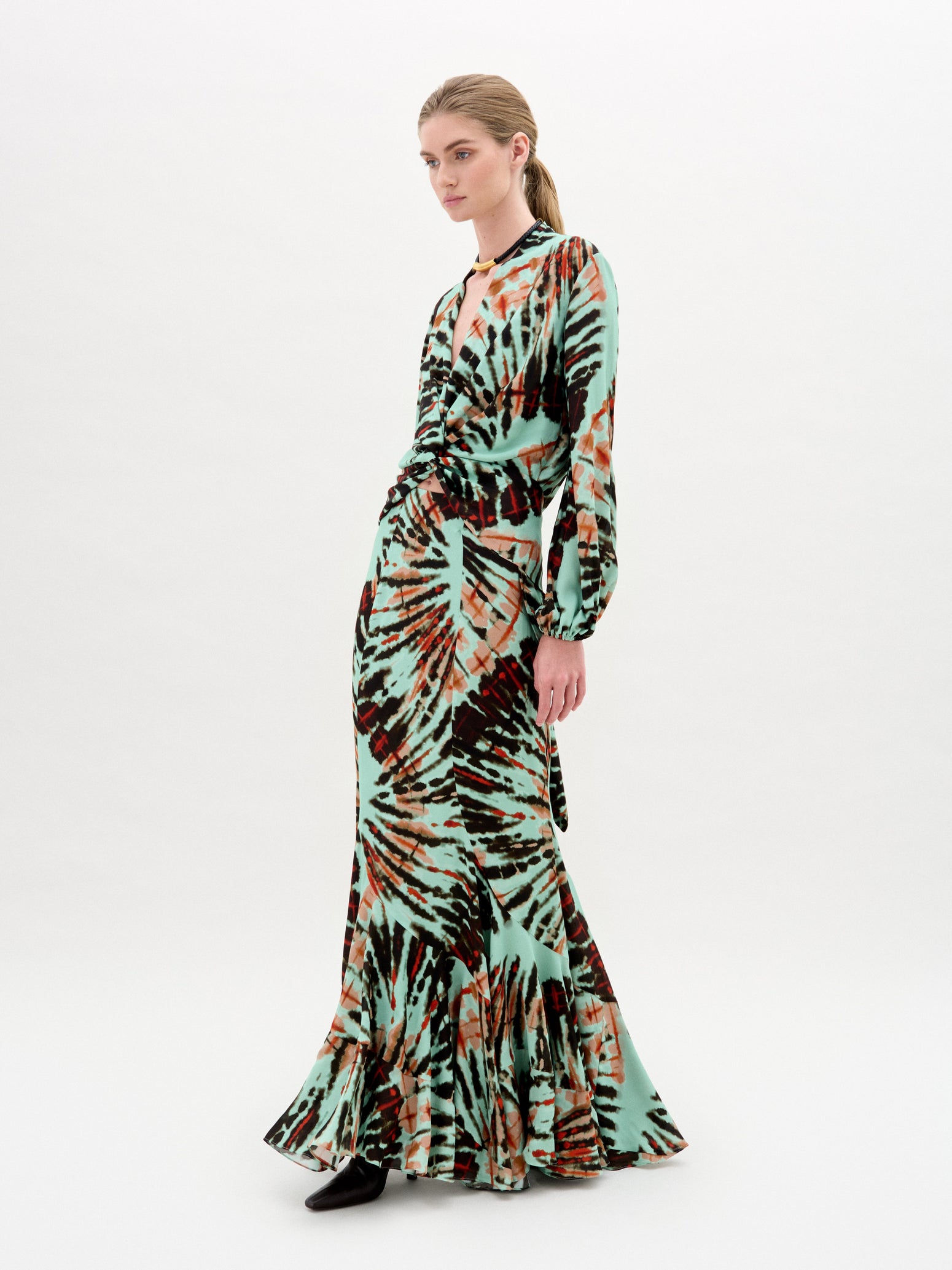 Against a plain white background, a person wears the Florina Skirt, crafted from luxurious Italian fabric. This long skirt showcases a vibrant tie-dye pattern with green, orange, and black hues cascading elegantly down its maxi length.