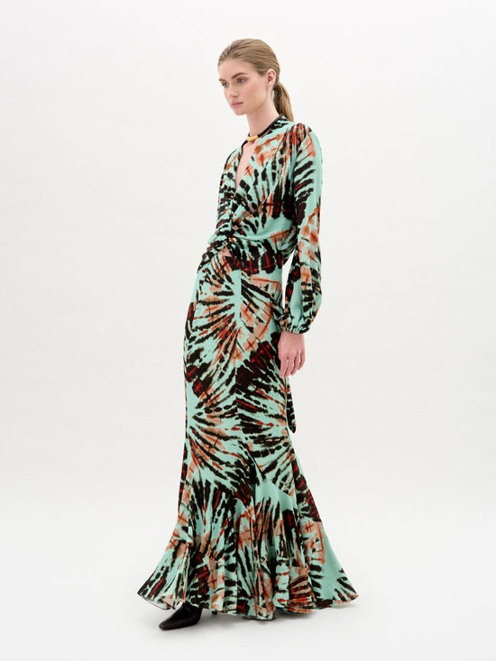 Against a plain white background, a person wears the Florina Skirt, crafted from luxurious Italian fabric. This long skirt showcases a vibrant tie-dye pattern with green, orange, and black hues cascading elegantly down its maxi length.