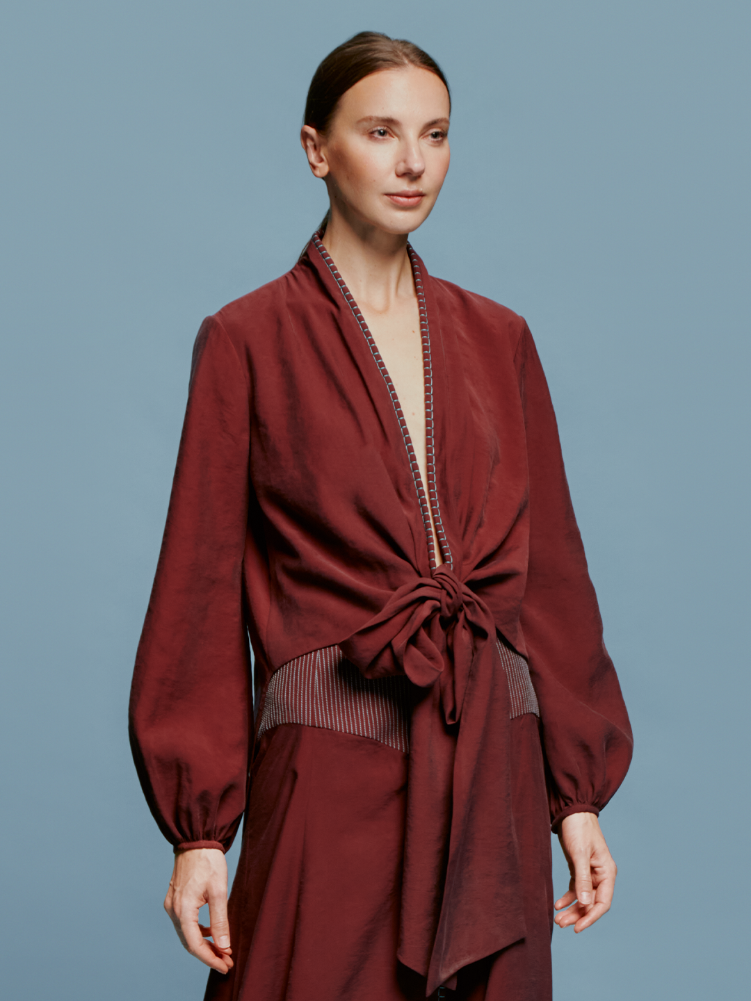 A woman with brown hair is wearing the Honey Blouse Burgundy, a wrap-style top with balloon sleeves, standing against a blue background, ready for the September 2024 launch.