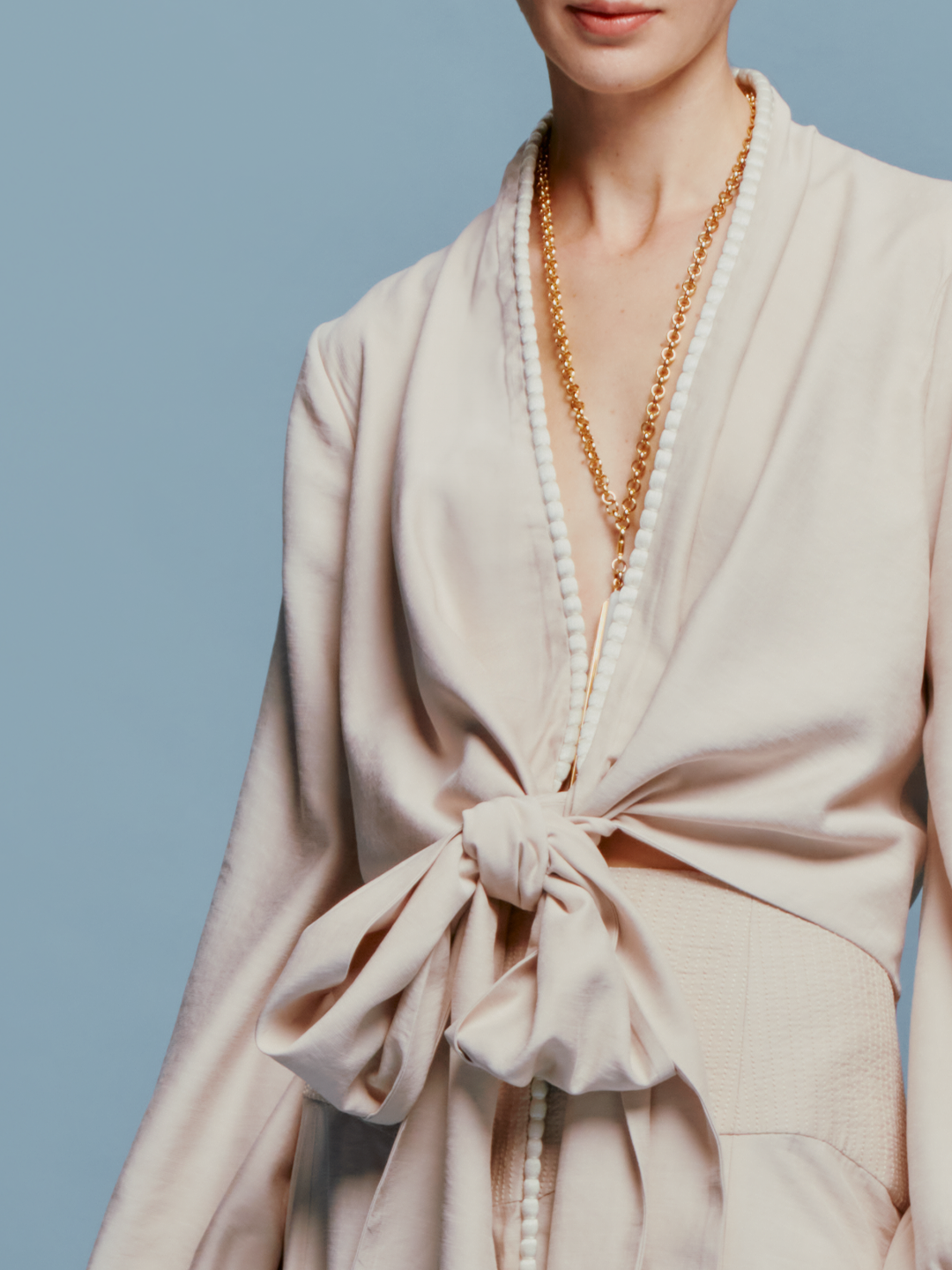 Person in a Honey Blouse Cream with a bow, wearing a long necklace, stands against a plain blue background, marking the date as either August 19th, 2024, or September 2nd.