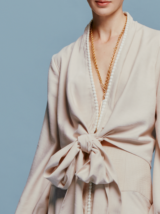 Person in a Honey Blouse Cream with a bow, wearing a long necklace, stands against a plain blue background, marking the date as either August 19th, 2024, or September 2nd.