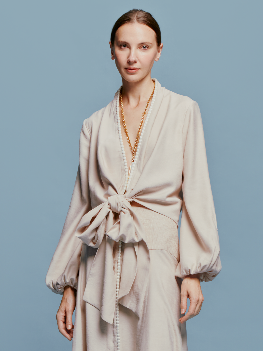 Person in a Honey Blouse Cream with a bow, wearing a long necklace, stands against a plain blue background, marking the date as either August 19th, 2024, or September 2nd.