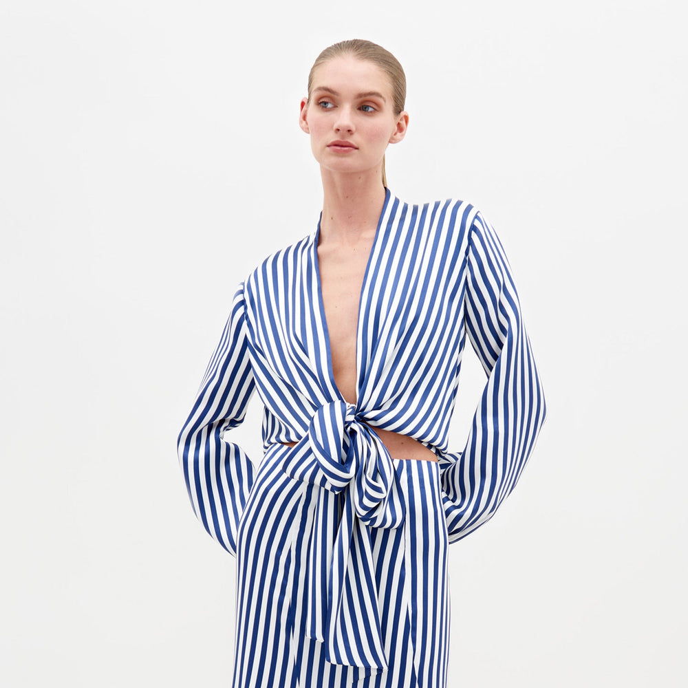 A person stands in the Honey Blouse White Blue Stripes, featuring a tie at the waist and a ruffled hem, paired with embellished sandals. This stylish ensemble is available for pre-order with shipping by February 15th.