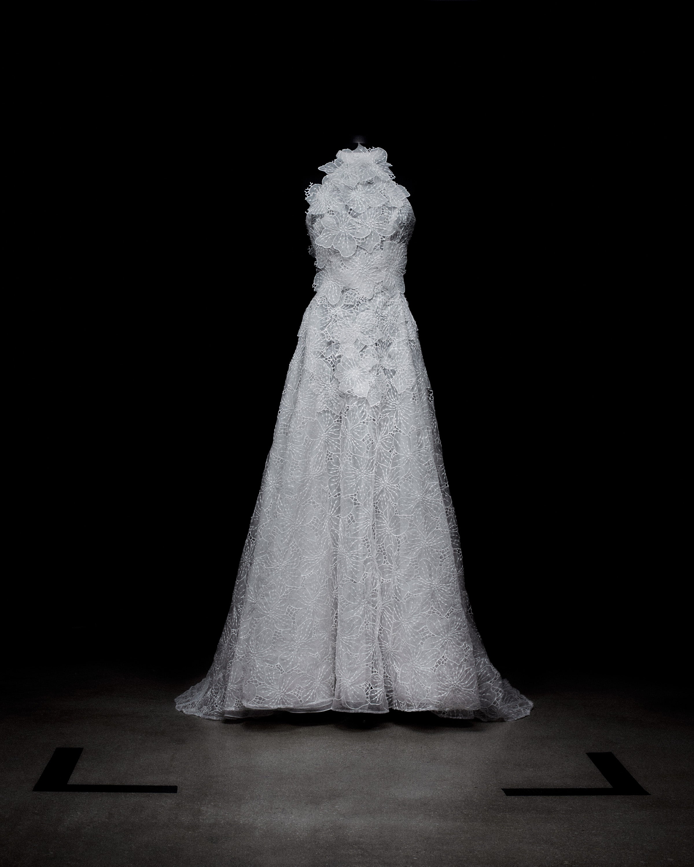 A white, intricately detailed lace gown is displayed against a black background on a mannequin.