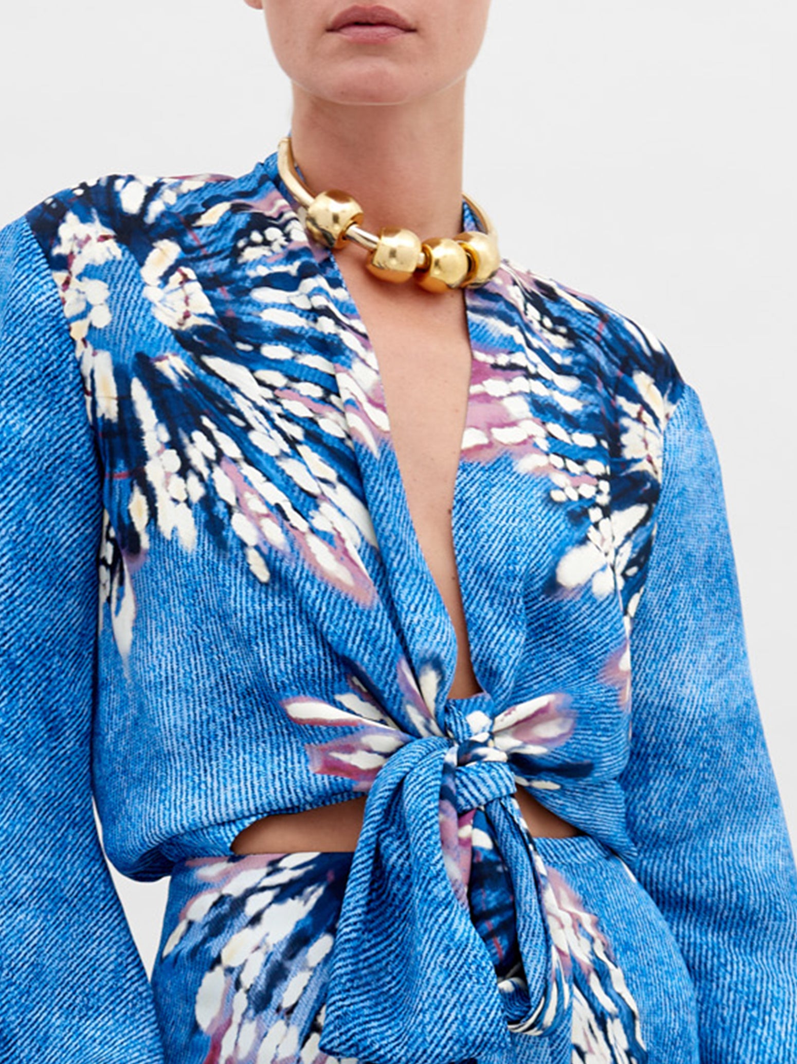 A woman in a blue floral outfit with balloon sleeves and a front tie stands against a plain background, wearing a gold beaded necklace, ready for pre-order until November 15th, 2024. The outfit she is wearing is the Honey Blouse Aciano Flower.