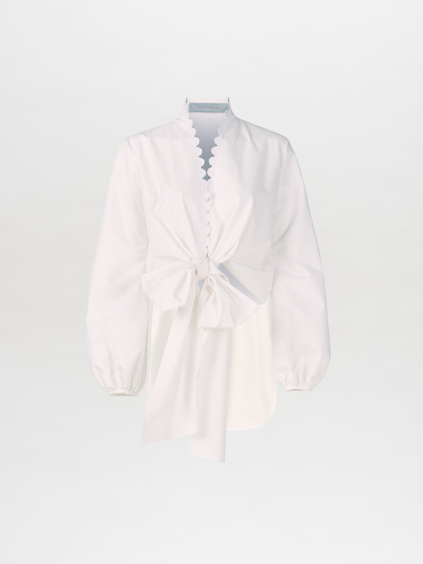 The Honey Blouse White Eyelet showcases a long-sleeve design in white, complete with scalloped edges, a button-up front, and a distinctive neckline highlighted by a large front bow. Made from soft cotton, this blouse seamlessly combines elegance with comfort.